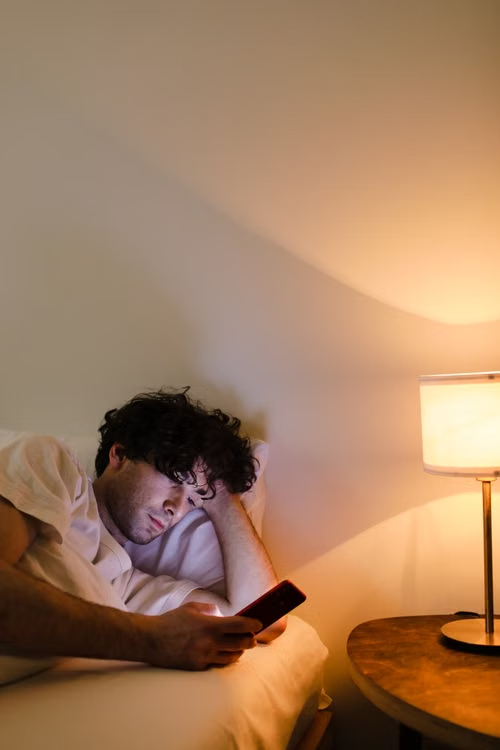 Man lying in bed scrolling on his phone next to a lit bedside lamp at night